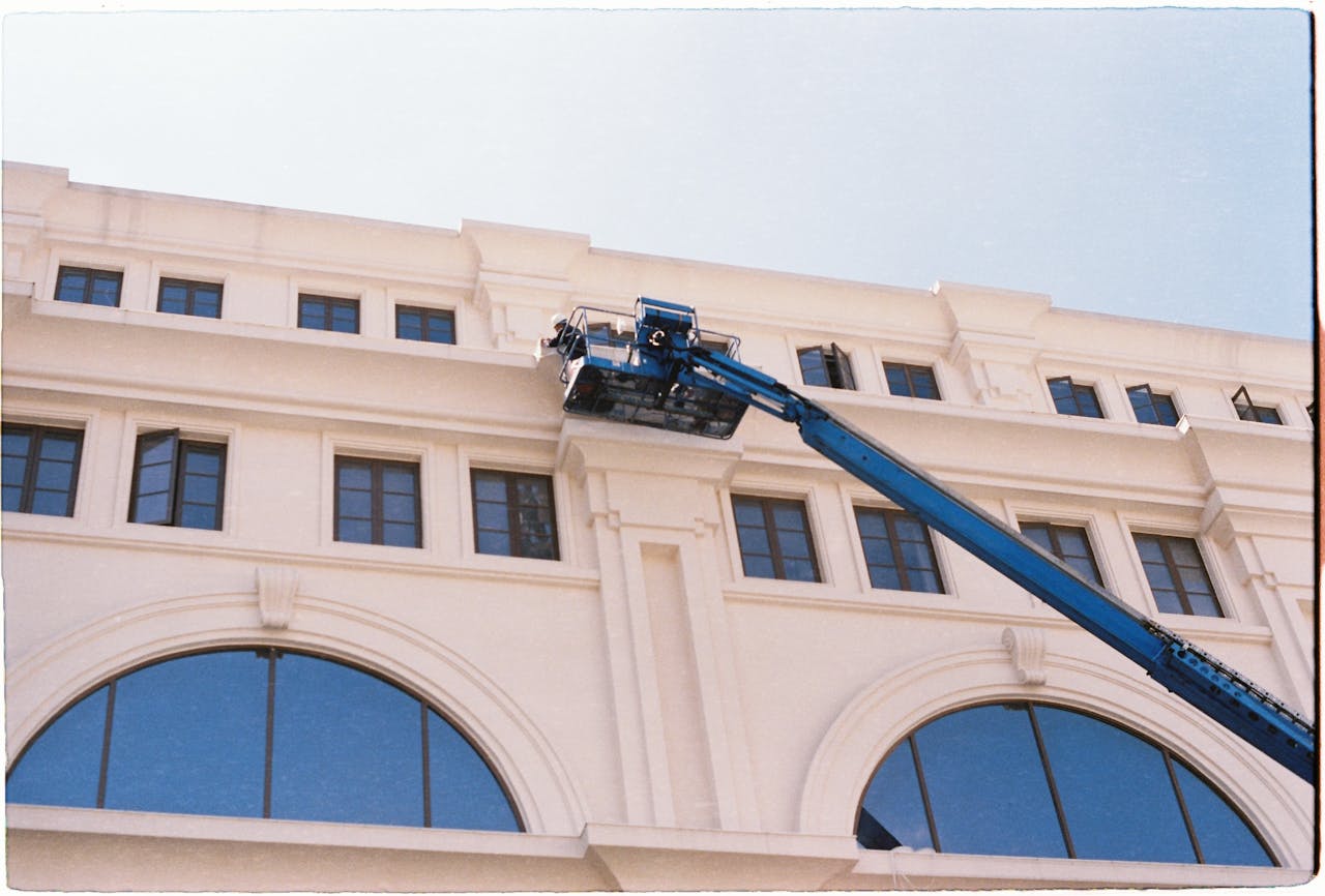 A worker cleans windows on a building using a blue crane on a clear day.
